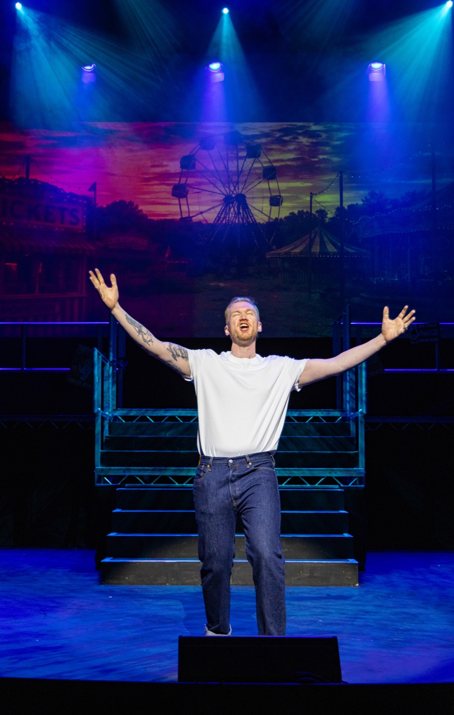 Jamie Edwardson performing on stage, arms outstretched in front of a fairground backdrop