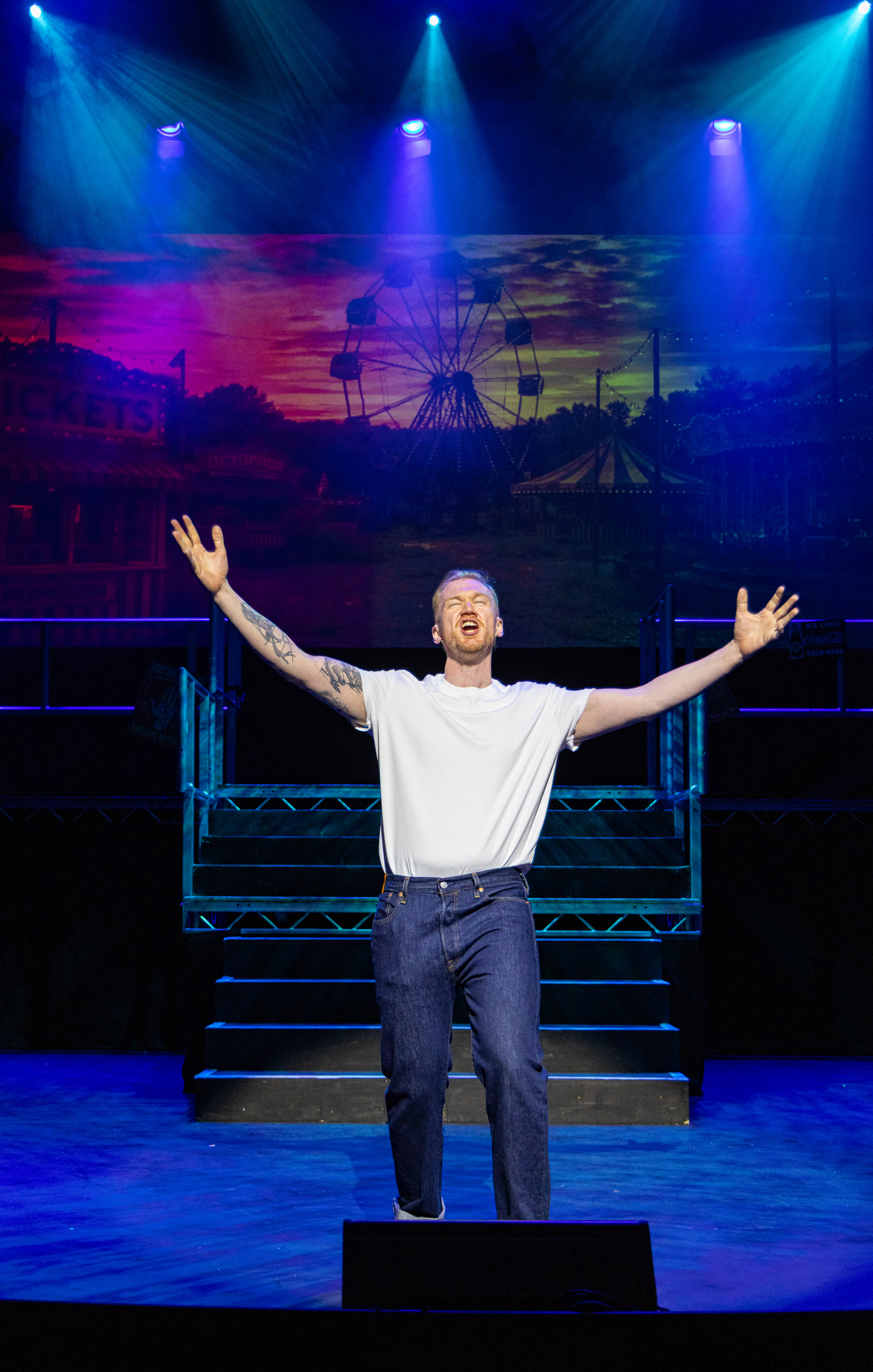 Jamie Edwardson performing on stage, arms outstretched in front of a fairground backdrop